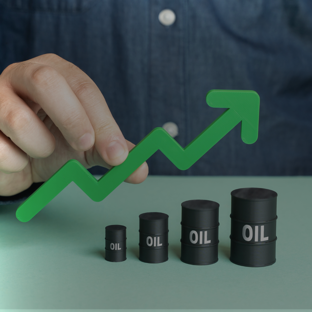A person’s hand holds a large, upward-trending green arrow above four black oil barrels of increasing size, symbolizing a sharp rise in crude oil prices.